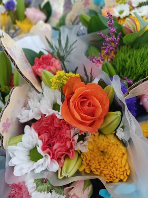 A close-up of a vibrant floral arrangement featuring an orange rose at the center, surrounded by white daisies with green centers, pink carnations, yellow chrysanthemums, and purple lavender sprigs. T