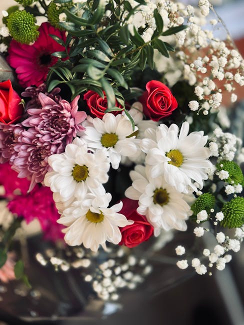 A vibrant floral bouquet featuring white daisies with central yellow discs, deep pink roses, and purple chrysanthemum blossoms with layered petals. The arrangement is accented with sprigs of delicate 