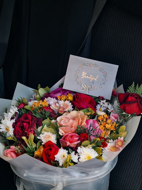 A vibrant flower bouquet arranged with a variety of fresh flowers including red roses, pink roses, light purple carnations, white daisies, and yellow filler flowers, all wrapped in white tissue paper.