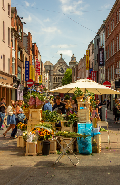 A lively outdoor scene on Brentwood High Street featuring a flower stall operated by flowersbrentwood.co.uk, displaying an array of fresh floral arrangements and bouquets. The stall is shaded by a lar