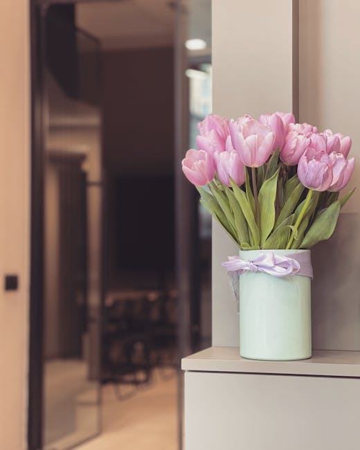 A elegant bouquet of light pink tulips with fresh green stems and leaves arranged in a pale green ceramic vase decorated with a lavender ribbon bow. The bouquet is placed on a white surface, likely a 