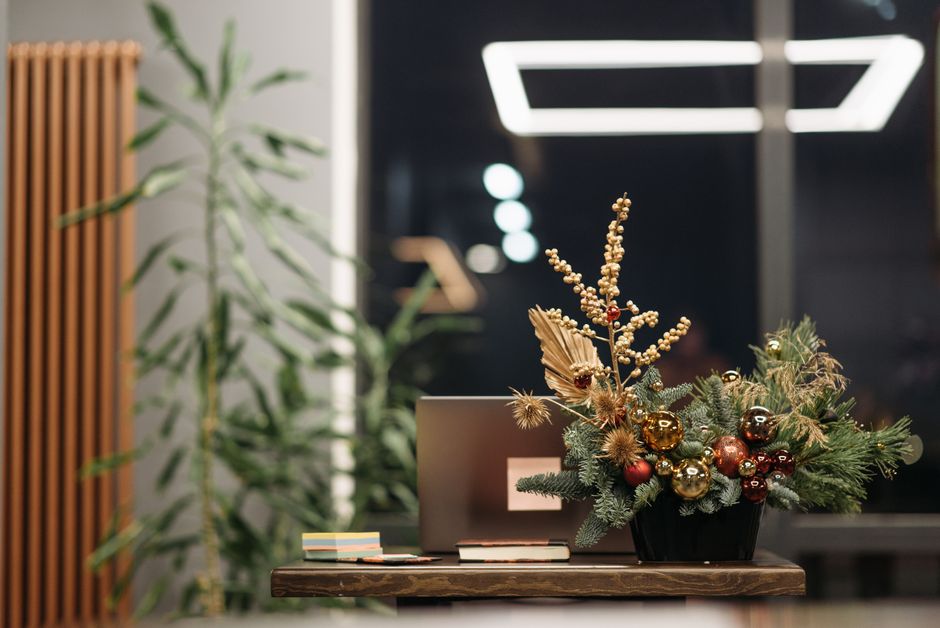 A festive floral arrangement featuring gold, bronze, and deep red Christmas baubles nestled among lush green pine branches, gold-tipped dried thistles, and golden berries, all arranged in a black vase